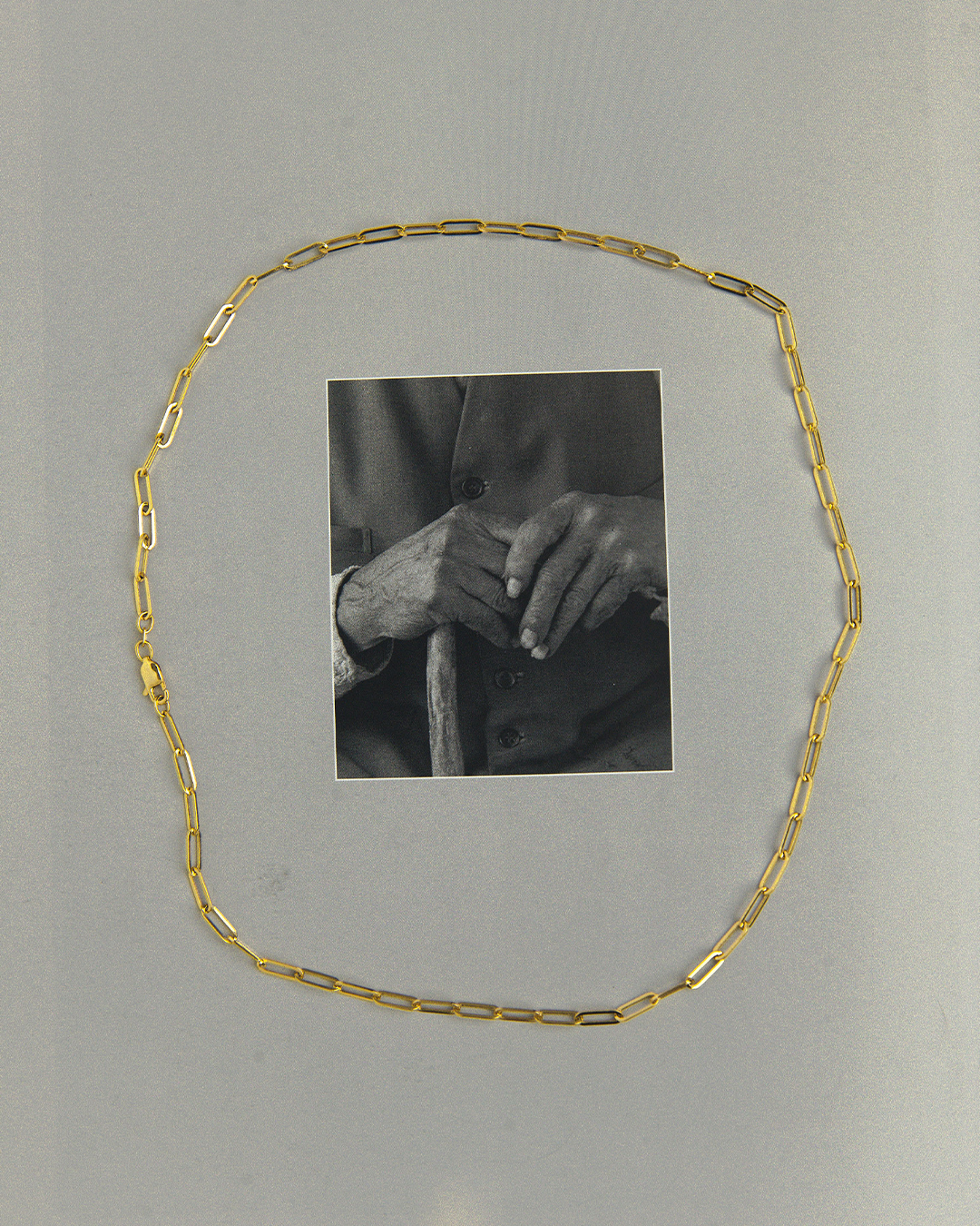 Cadena tipo clip de oro amarillo de 14 quilates dispuesta sobre un fondo gris, enmarcando una fotografía en blanco y negro de unas manos, con énfasis en el diseño de sus eslabones alargados y el cierre de brochE.