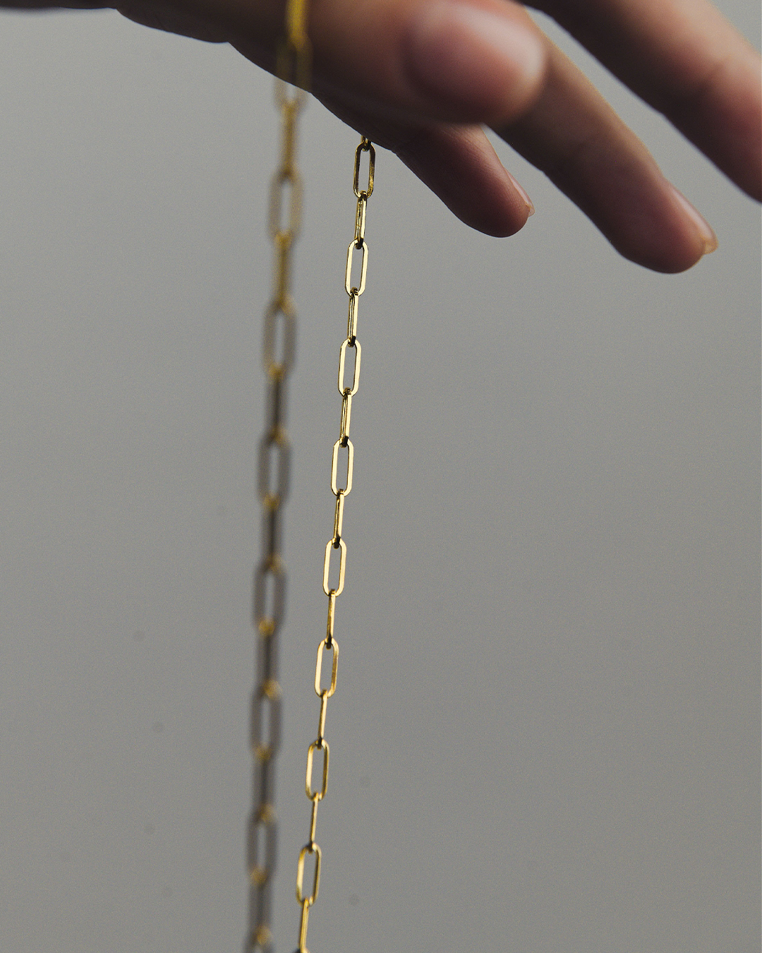 Detalle de una cadena de M18 tipo clip de oro amarillo de 14 quilates sostenida entre los dedos de un modelo, destacando el diseño de eslabones alargados y el acabado pulido sobre un fondo neutro desenfocado.