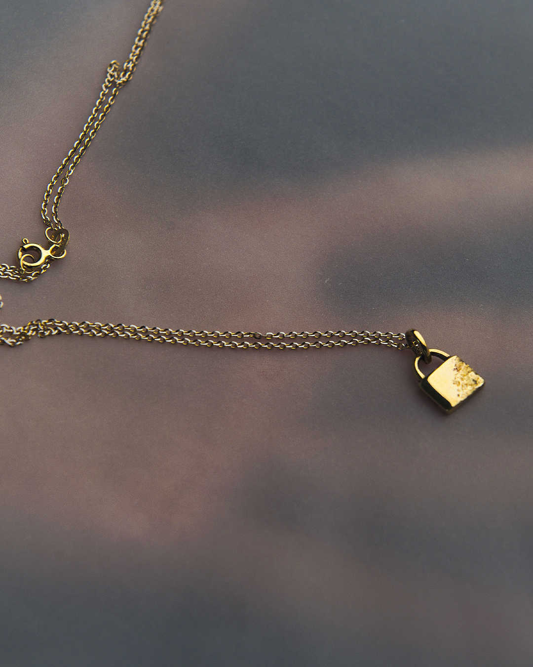 Cadena de oro con dije en forma de candado de oro amarillo de 10 quilates marca M18. Sobre una superficie lisa, mostrando el acabado pulido y el diseño minimalista del colgante.
